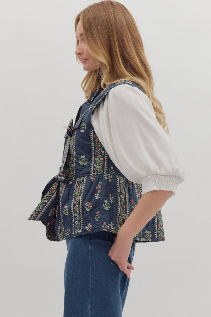 Woman wearing a floral patterned vest over a white shirt and blue jeans on a light gray background
