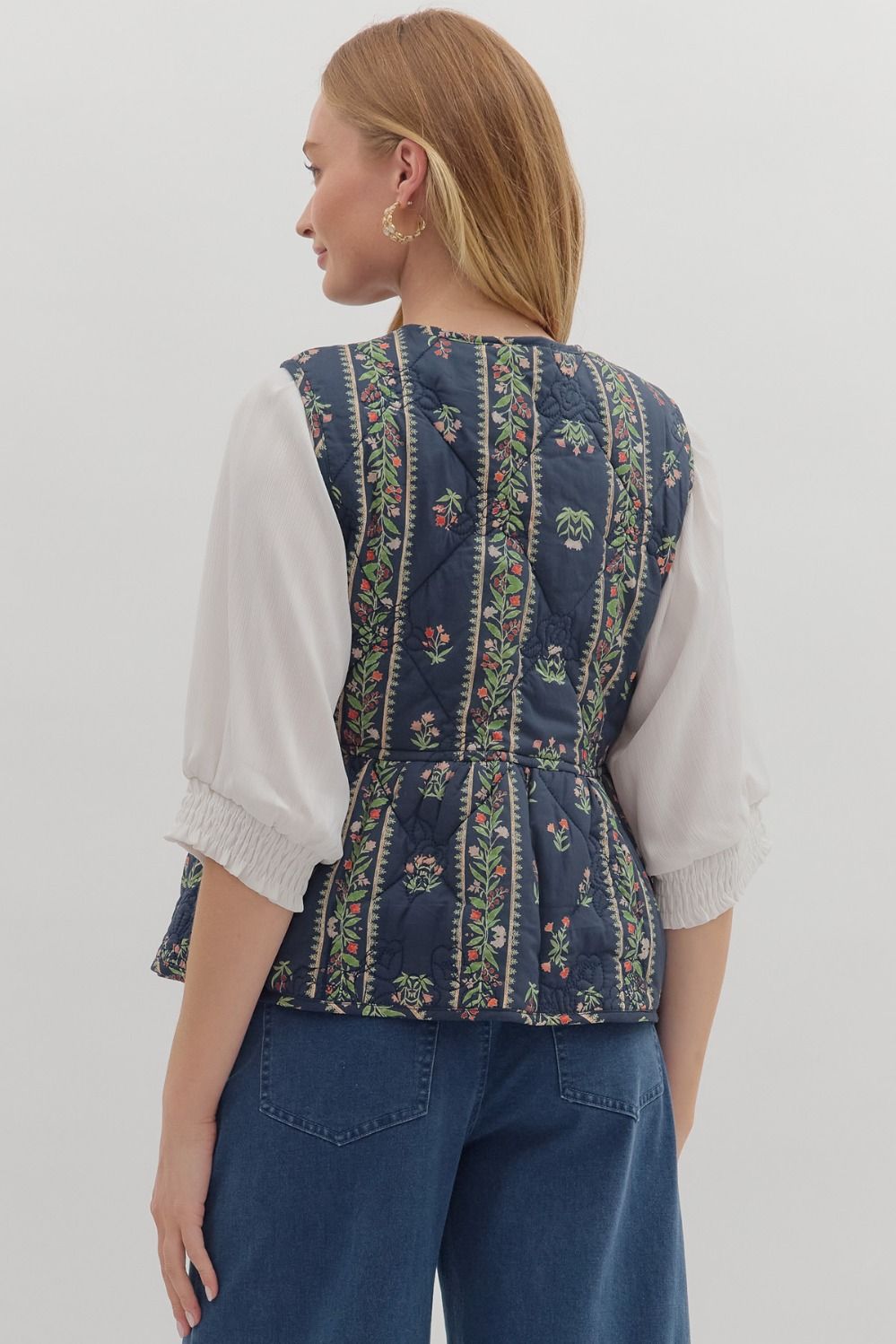 Person wearing a blue patterned vest over a white shirt and blue jeans on a light gray background