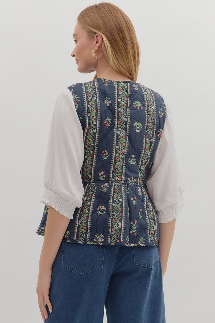 Person wearing a blue patterned vest over a white shirt and blue jeans on a light gray background